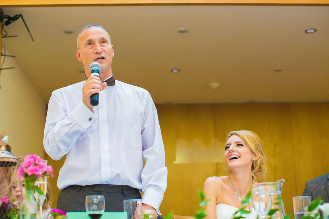 A man in a tuxedo, jacket removed, is speaking into a microphone, giving a father of the bride speech. The bride is seated to his left, and is laughing.