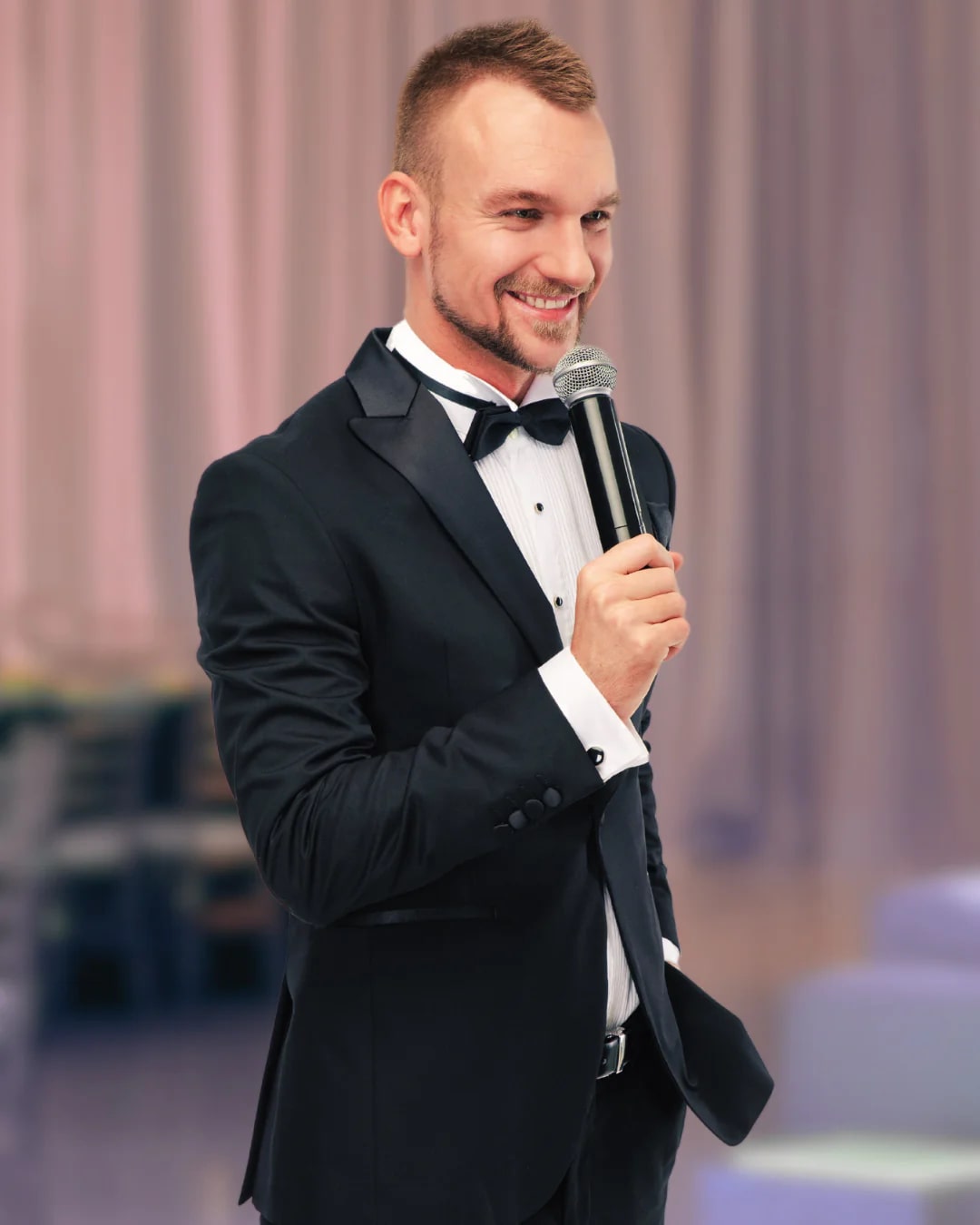 A handsome man in a tuxedo smiles and speaks into a microphone, delivering a funny best man speech