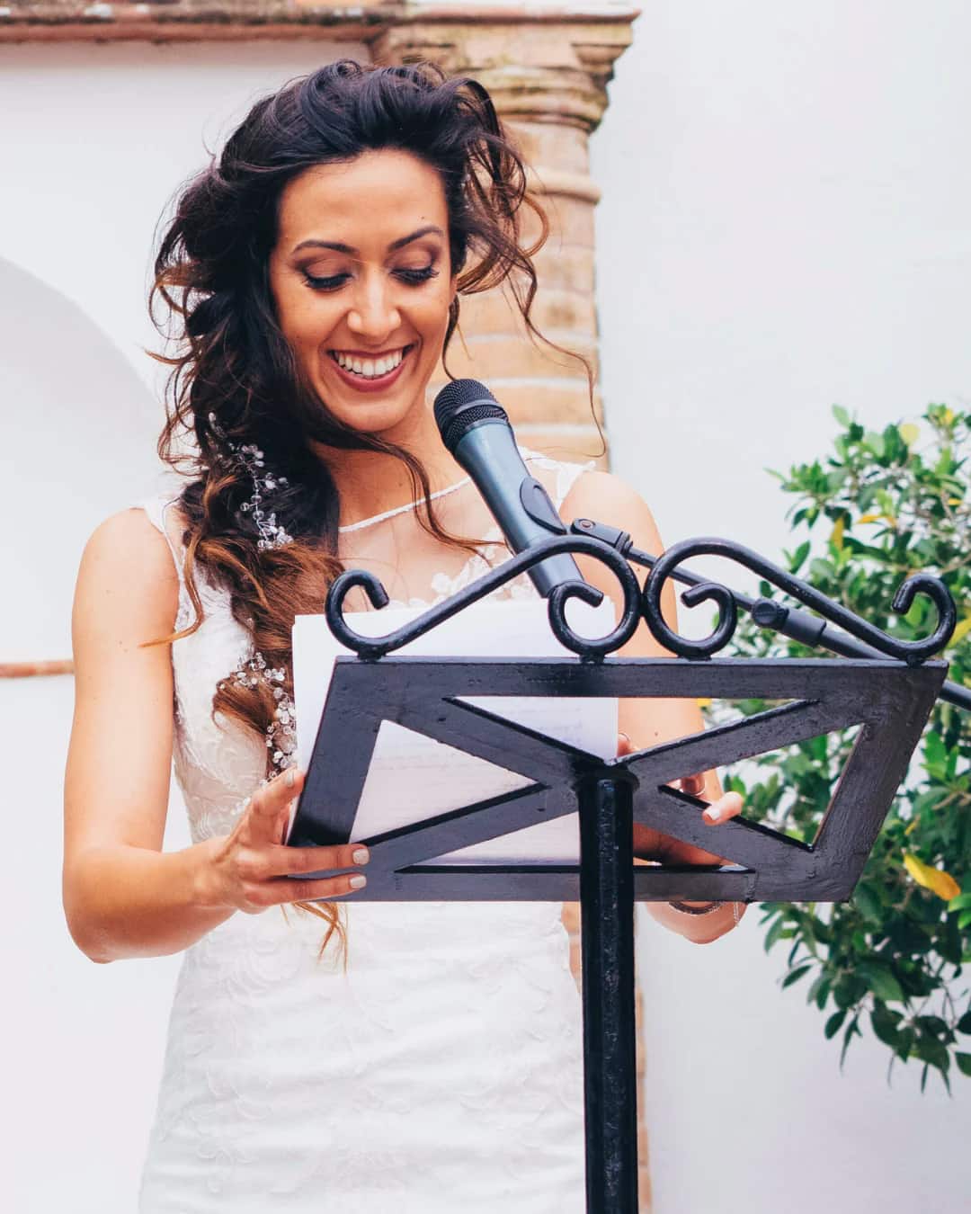 A young woman with long, dark hair, wearing an off white bridesmaid dress stands at a podium with notes in front of her. She is smiling and speaking into a microphone giving a maid of honor wedding speech.