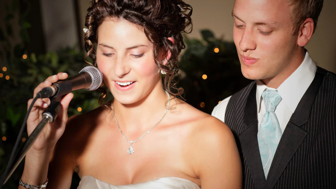 A bride speaks into a microphone, giving a wedding thank you speech, as the groom smiles at her.