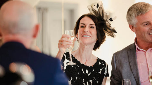 An older woman, likely the mother of the bride or mother of the groom, wears a stylish black dress and holds up a glass of champagne, giving a wedding toast or wedding speech.